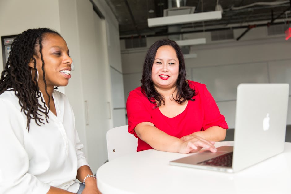 Two women engaging in a collaborative discussion in a modern office setting, using a laptop.